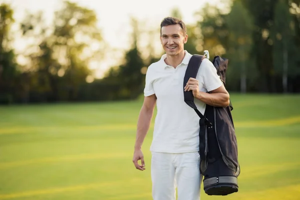 Um homem de fato branco anda pelo campo de golfe com um saco de golfe e sorri. — Fotografia de Stock