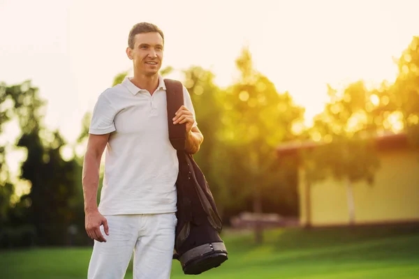 Un hombre con un traje blanco camina por el campo de golf con una bolsa de palos de golf contra una hermosa puesta de sol —  Fotos de Stock