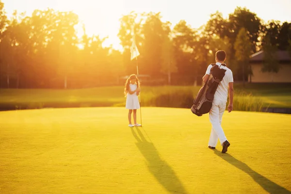 En man i vit kostym och en golfklubb väska går mot flickan som står bredvid hålet i solnedgången — Stockfoto