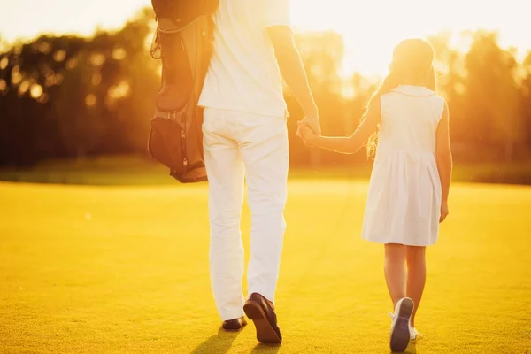 Padre e hija están caminando a lo largo del campo de golf contra el fondo del atardecer tomados de la mano . — Foto de Stock