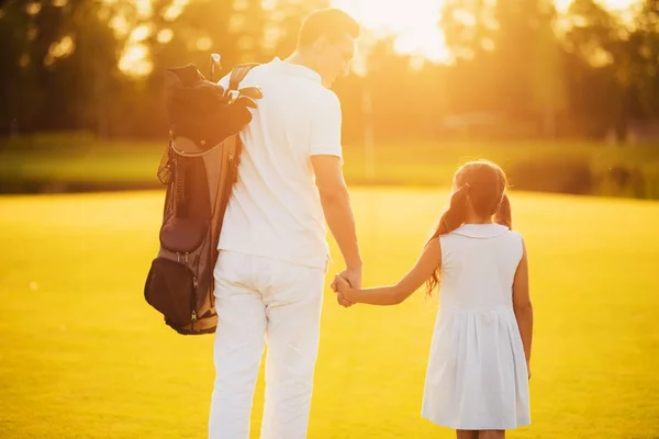 Uomo con una borsa per mazze da golf e una ragazza che cammina lungo un campo da golf tenendosi per mano su uno sfondo al tramonto — Foto Stock