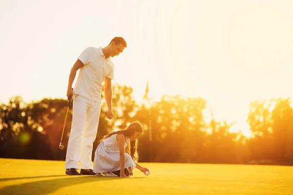 Um homem ensina uma rapariga a jogar golfe. menina coloca uma bola em um suporte, homem segura um pau — Fotografia de Stock
