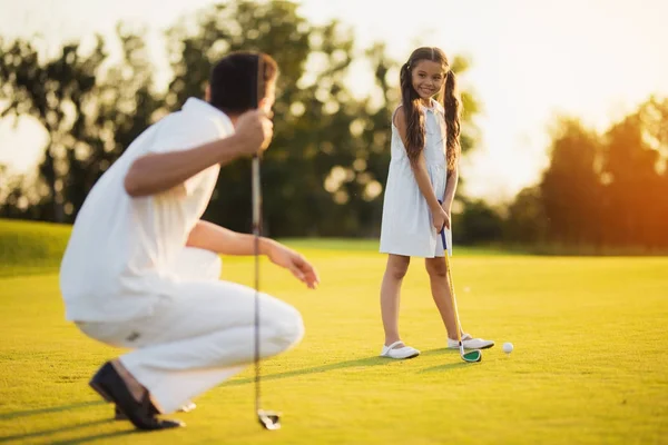 Otec dřepy golfovou holí v ruce a se dívá na jeho dceru, která se dívá na něj a připravuje se trefit míč — Stock fotografie