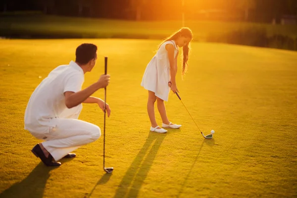 Chica jugando al golf sobre un fondo al atardecer. El hombre se agacha junto a ella y se apoya en un palo de golf — Foto de Stock