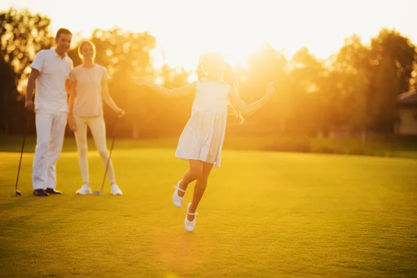 The girl is running against the sunset. Her parents are standing with golf clubs in their hands behind her — Stock Photo, Image