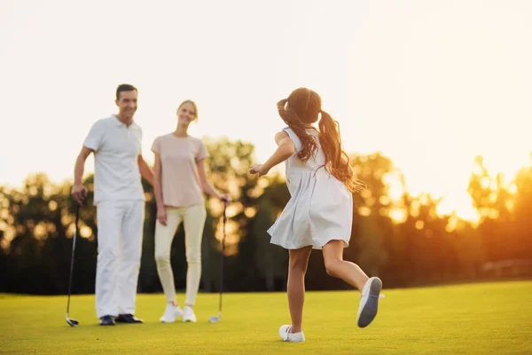A menina corre através do campo de golfe em direção ao homem e mulher que estão na frente dela e tacos de golfe — Fotografia de Stock