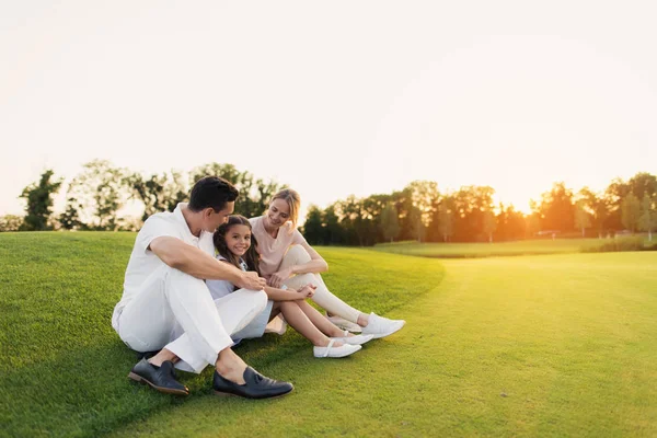 Família feliz descansando em um campo de golfe sentado na grama em um fundo pôr do sol — Fotografia de Stock