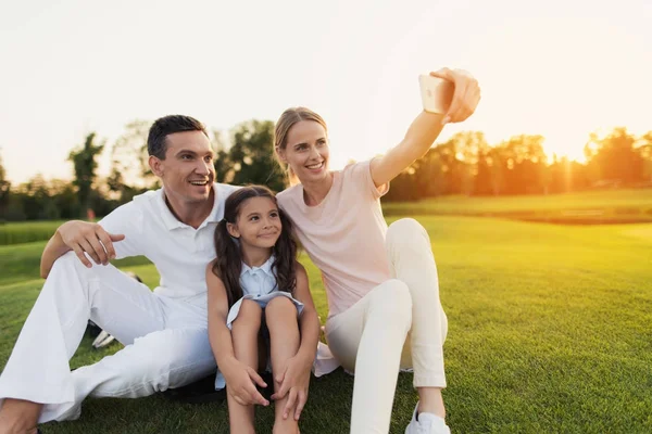Famiglia facendo selfie su smartphone grigio seduto sull'erba sul campo da golf sullo sfondo del tramonto — Foto Stock