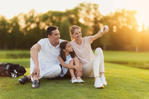 Una mujer, un hombre y una chica que se sientan entre ellos, se sientan en un campo de golf y hacen selfie en un teléfono inteligente gris — Foto de Stock