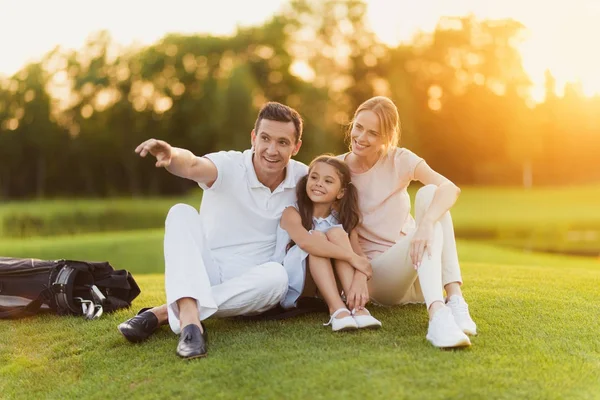 A família senta-se na grama no campo de golfe, o homem aponta para o lado, ea esposa e filha olhar lá — Fotografia de Stock