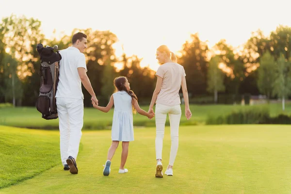 La familia está caminando a lo largo del campo de golf. Madre y padre tomados de la mano de su hija — Foto de Stock