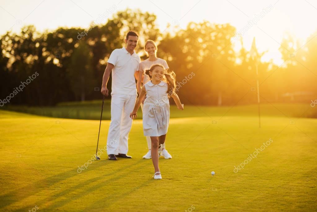 Happy family posing on a golf course on a sunset background. The girl smiles and runs towards the camera