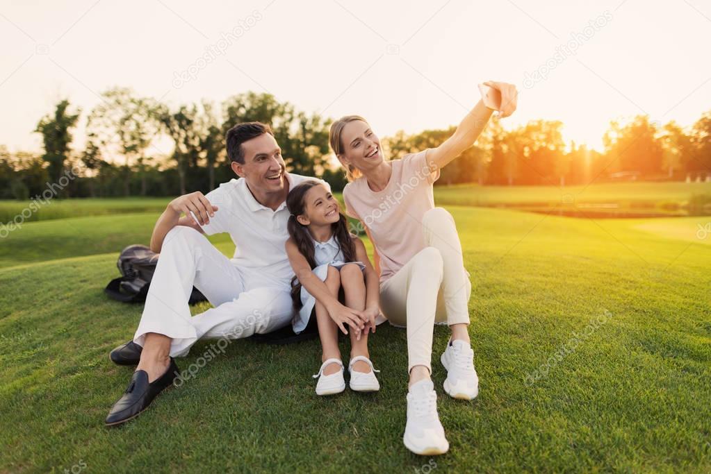 Happy family sitting on grass at golf course and posing on smartphone, making selfie