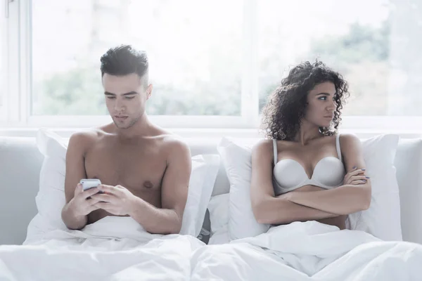 A young couple is sitting on the bed. The guy is looking at something on his smartphone. The girl is offended by him