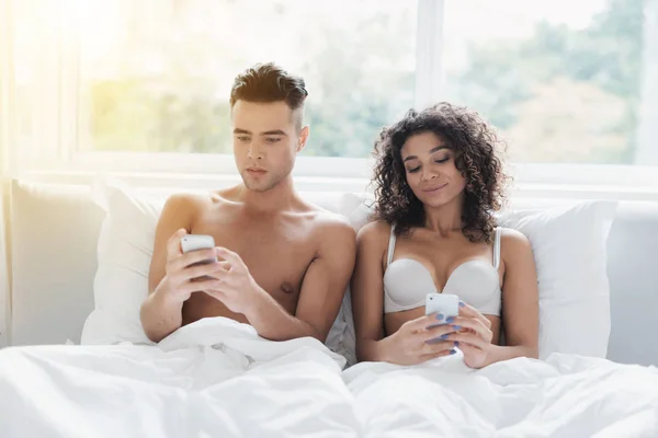 Una joven pareja está sentada en la cama. un chico y una chica están viendo algo en sus teléfonos inteligentes . — Foto de Stock