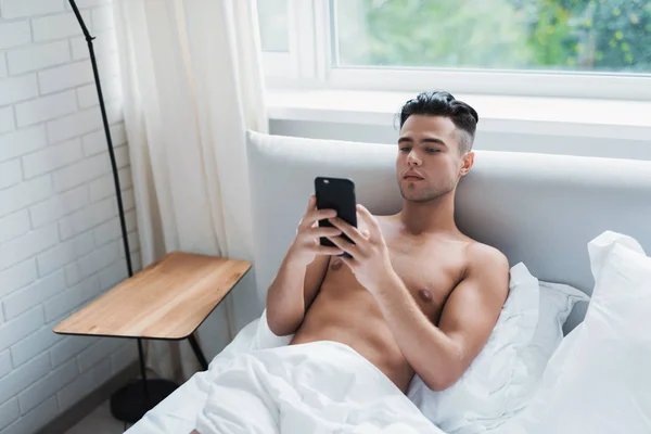 Facet leży w białym łóżku i wygląda na ekranie jego czarny smartphone. — Zdjęcie stockowe