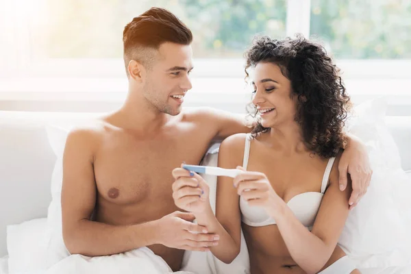 Νεαρό ζευγάρι κάθεται στο κρεβάτι και κοιτάζοντας κοπέλα τεστ εγκυμοσύνης. Είναι χαρούμενος και χαμογελαστός. — Φωτογραφία Αρχείου