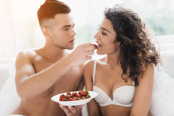 Pria dan gadis itu duduk di tempat tidur dengan pakaian dalam mereka. Mereka makan secara seksual satu sama lain dengan stroberi . — Stok Foto