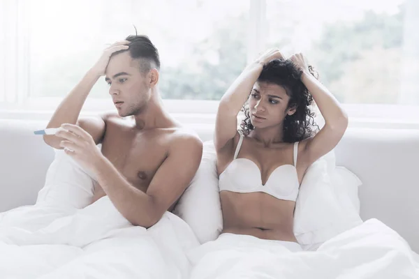 Young couple sitting on bed in underwear. They have a pregnancy test and they look unhappy.