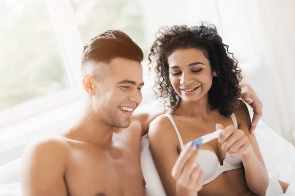 Young couple sitting on bed and looking at pregnancy test girl. They are happy and smiling.