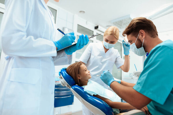 A dentist is preparing to treat the teeth of a little girl. There are nurses around. Girl smiling