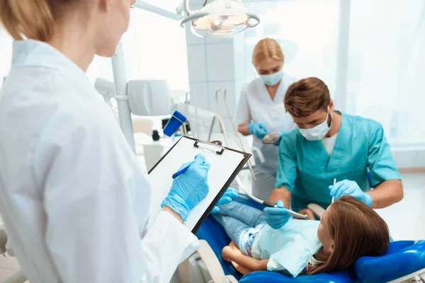 Eine Krankenschwester schreibt die Handlungen eines Zahnarztes in einem medizinischen Journal auf, während er ein Mädchen behandelt — Stockfoto