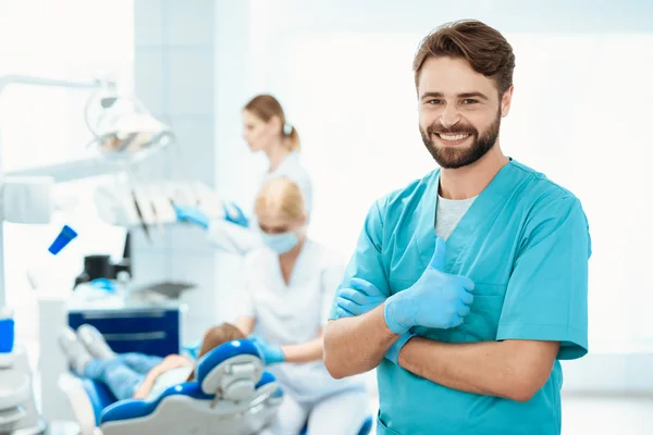 Un dentista posa sobre el telón de fondo de un consultorio dental. Muestra los pulgares hacia arriba —  Fotos de Stock