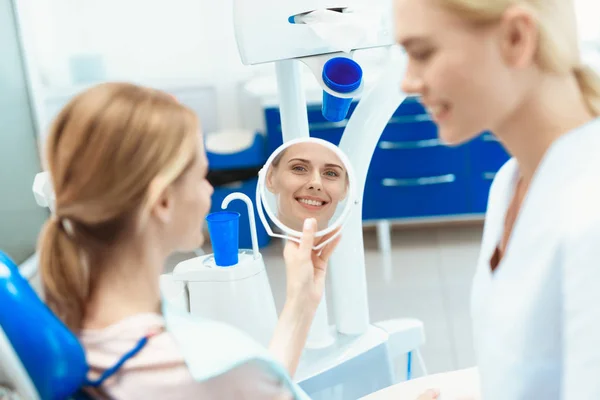 Een vrouw is in een tandheelkundige stoel zitten en in de spiegel kijken. Er is een tandarts in de buurt — Stockfoto