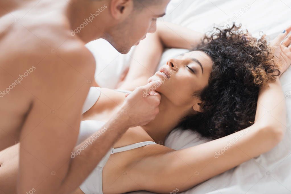 A young couple in a sexual pose is lying on a white bed. The guy is over the girl. He gently caresses her. They are in a bright bedroom.