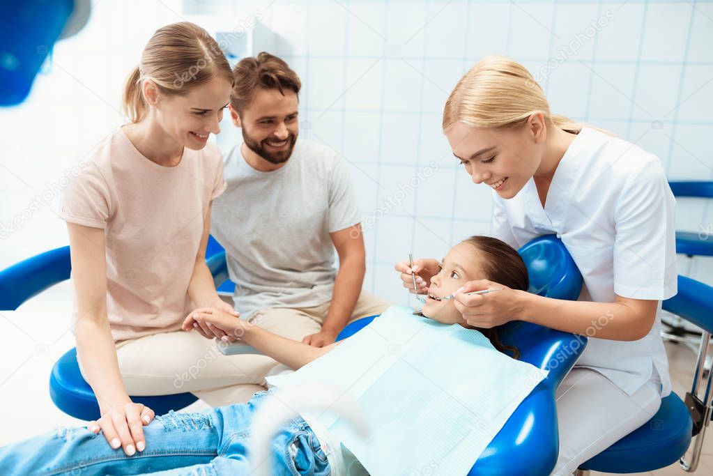 Parents brought their daughter to see a dentist. The girl is sitting in the dental chair. A doctor stands nearby