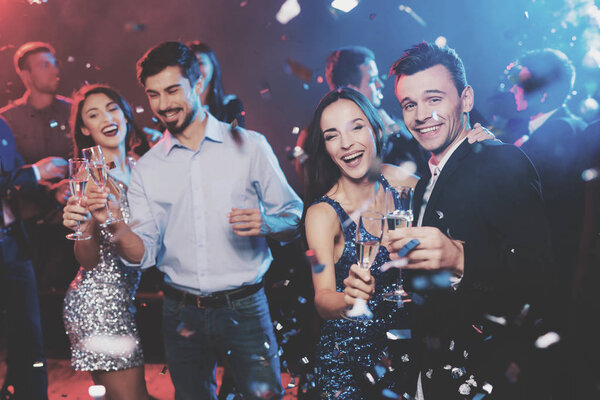 Young people have fun at a New Year's party. In the foreground two pairs dance. The girls are wearing gray and blue dresses in sequins. On one of the guys a light shirt, on the other a black jacket.