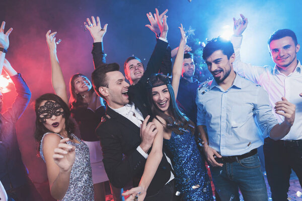 New Year party. Young couple dancing with glasses of champagne in hands. Against the background, young people's friends are dancing. Around fly confetti and there is white smoke.