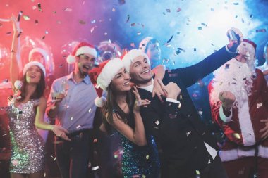 Genç bir çift yeni yıl partisinde eğleniyor. Onlar selfi bir smartphone olun ve dışarı çıktım. Çok eğlenceli. Onların arkadaşları dans ve çevresinde sinek konfeti arka planı.