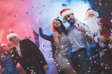Genç çift bardak şampanya elinde dans. Onların arkasında onların arkadaşları dans ve Noel Baba gibi giyinmiş bir adam. Renkli duman arka planı, konfeti etrafında sinekler.