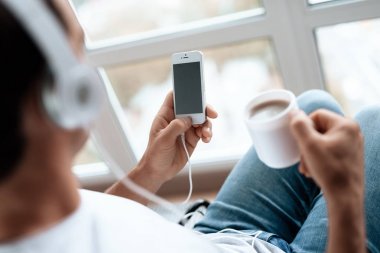 Bir özürlü tekerlekli büyük bir panoramik pencere önünde oturur ve Smartphone müzik dinler.