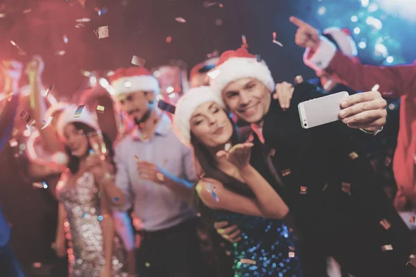 Young couple having fun at New Year's party. They make selfi on a smartphone and go out. They are very fun. Against the background of dancing their friends and around fly confetti.