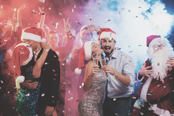 Casal jovem dançando com copos de champanhe nas mãos. Atrás deles dançam seus amigos e homens vestidos de Papai Noel . — Fotografia de Stock