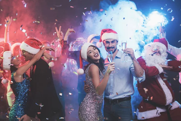 Casal jovem dançando com copos de champanhe nas mãos. Atrás deles dançam seus amigos e homens vestidos de Papai Noel . — Fotografia de Stock