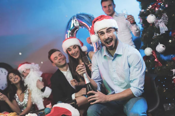 Um homem de chapéu de Pai Natal abre uma garrafa de champanhe. Os amigos de um homem sentam-se ao lado dele e um cara vestido de Papai Noel — Fotografia de Stock