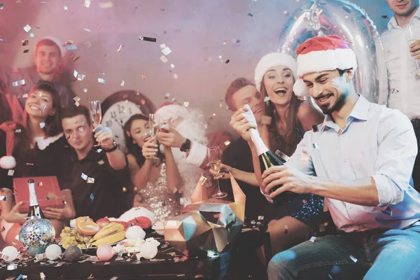 Un homme au chapeau de Père Noël ouvre une bouteille de champagne. Les amis d'un homme s'assoient à côté de lui et un gars habillé en Père Noël — Photo