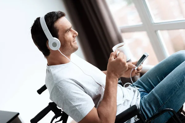 A disabled person in a wheelchair sits in front of a large panoramic window and listens to music on the smartphone. He has a smartphone in his hands and headphones on his head.