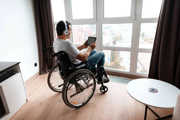 A pessoa com deficiência em uma cadeira de rodas se senta na frente de uma grande janela panorâmica e olha para algo em seu tablet . — Fotografia de Stock