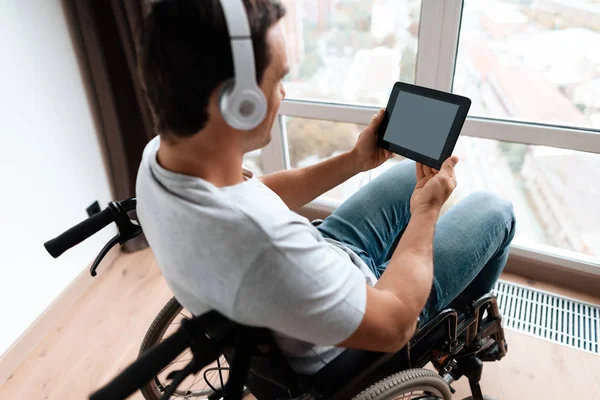 車椅子の障害者の大パノラマの窓の前に座って、彼のタブレット上の何かを見る. — ストック写真