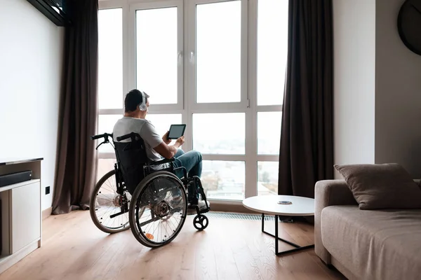 車椅子の障害者の大パノラマの窓の前に座って、彼のタブレット上の何かを見る. — ストック写真