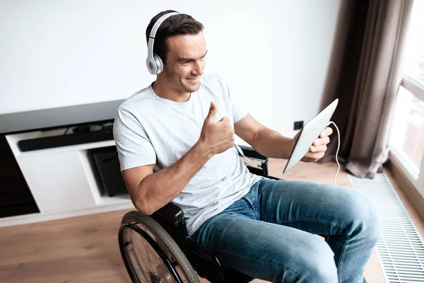 En funktionshindrad person i rullstol sitter framför stora panoramafönster och kommunicerar via video via sin surfplatta. — Stockfoto