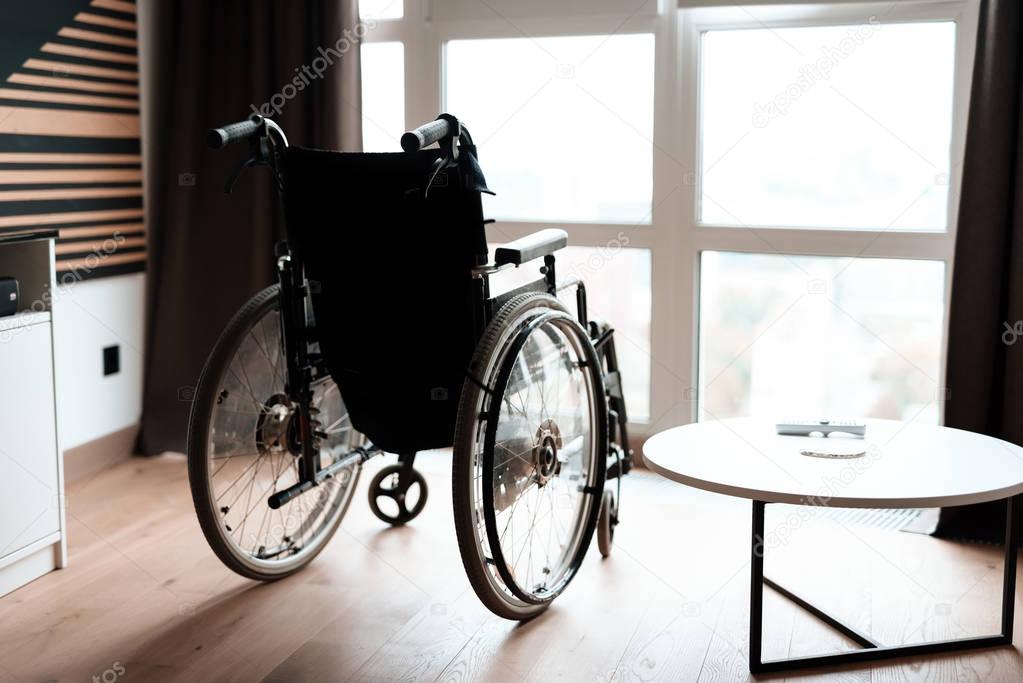 A modern wheelchair stands on the background of a large panoramic window in a modern bright living room.