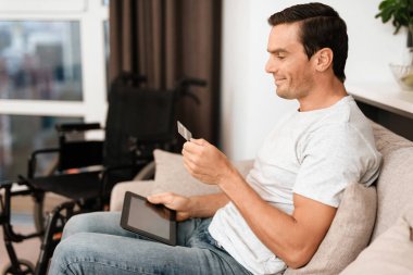 Engelli kişi koltukta oturuyor. O elinde bir tablet ve bir kredi kartı var.