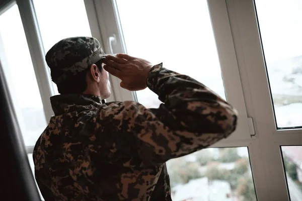 A man in uniform wears a large panoramic window and salutes. He has a serious face and he looks into the distance.