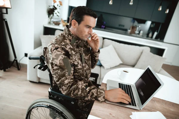 Discapacitado en uniforme militar, trabaja para su portátil. Se sienta en su escritorio. . — Foto de Stock