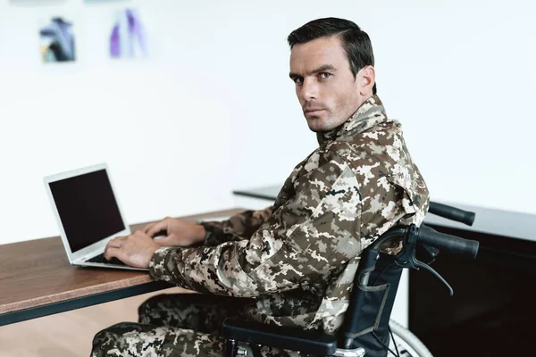A disabled military man is sitting at a table and working with a laptop. Behind it is a large panoramic window.
